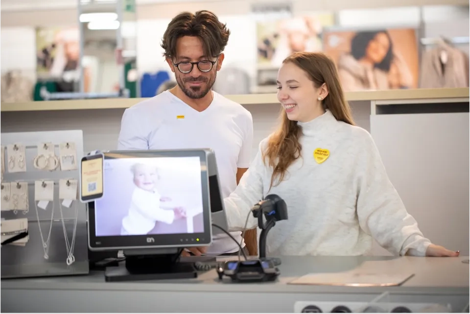 Ein Mann und eine Frau stehen lächelnd an der Kasse eines Ladens. Die Frau bedient die Kasse, auf einem Monitor ist das Foto eines lächelnden Babys zu sehen. Schmuck liegt auf der Theke.