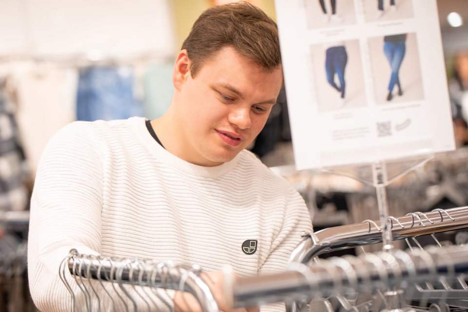 Ein Mann in einem weißen Pullover stöbert in einem Geschäft durch Kleiderständer und sucht nach Artikeln zum Kauf. Im Hintergrund ist eine Auslage mit Jeansbildern zu sehen.