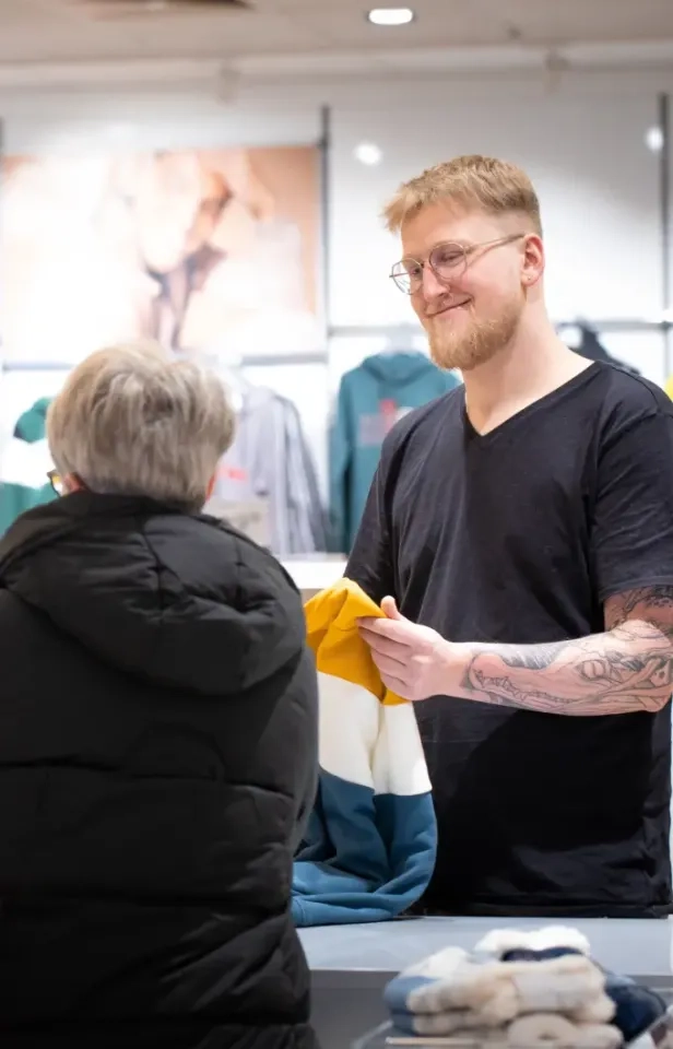Ein lächelnder Mann mit Brille und Tattoos reicht einer älteren Person im schwarzen Mantel an der Kasse eines Bekleidungsgeschäfts einen bunten, gefalteten Pullover. Im Hintergrund sind Kleiderständer und verschwommene Plakate zu sehen.