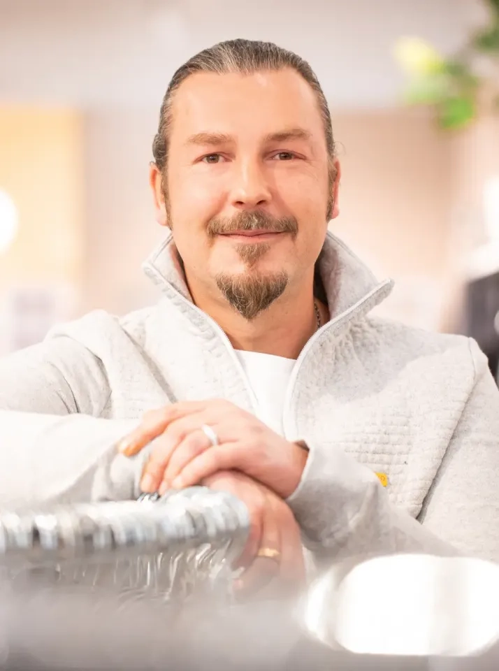 Ein Mann mit zurückgekämmtem Haar, Spitzbart und Schnurrbart, der einen hellgrauen Pullover mit Reißverschluss trägt, lächelt sanft, während er seine Hände auf einer Metalloberfläche in einem Innenbereich mit sanfter, verschwommener Beleuchtung ablegt.
