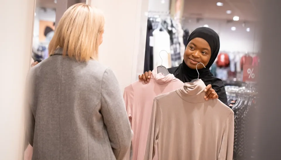 Eine Frau mit Hijab lächelt, während sie in einem Geschäft einer anderen Frau zwei Rollkragenoberteile hochhält, während sie die Kleiderauswahl besprechen und vergleichen.
