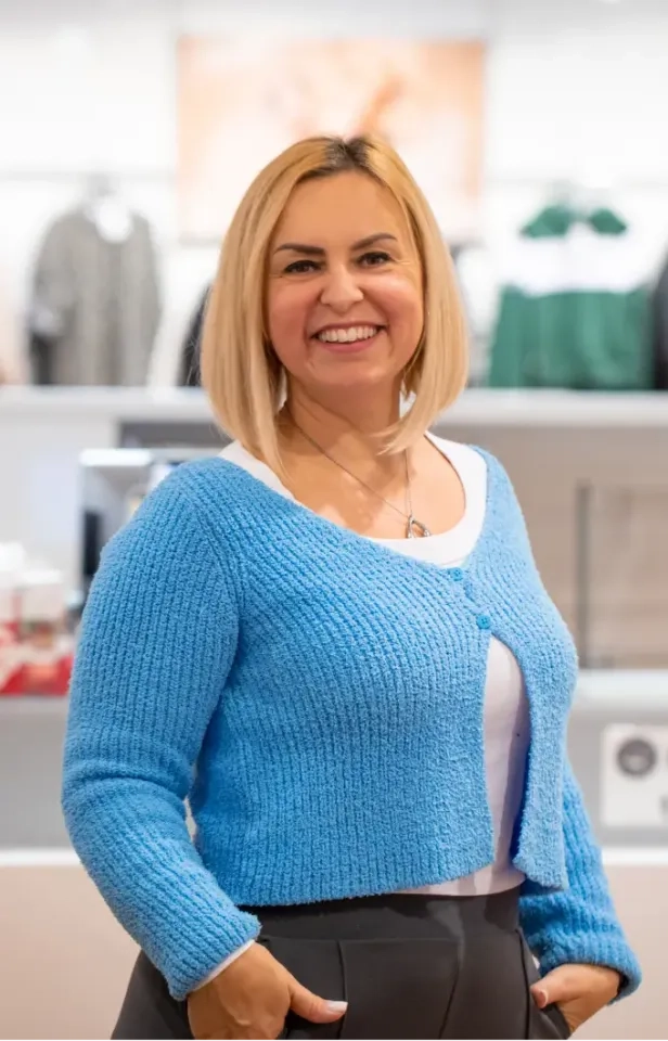 Eine lächelnde Frau mit glattem blondem Haar, die eine blaue Strickjacke über einem weißen Oberteil trägt, steht mit den Händen in den Taschen in einem Geschäft. Der Hintergrund ist leicht verschwommen und zeigt Regale und Kleidung in einem Geschäft.