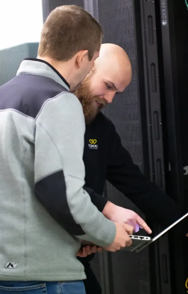 Zwei Personen stehen neben einem Serverschrank und unterhalten sich auf einem aufgeklappten Laptop. Eine Person zeigt auf den Bildschirm, die andere schaut aufmerksam zu.