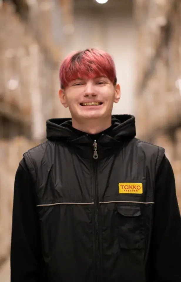 Eine lächelnde junge Person mit kurzen, rosa Haaren trägt eine schwarze Weste mit einem gelben Takko-Logo und steht in einem Lagergang, der mit Regalen voller Kartons gesäumt ist.