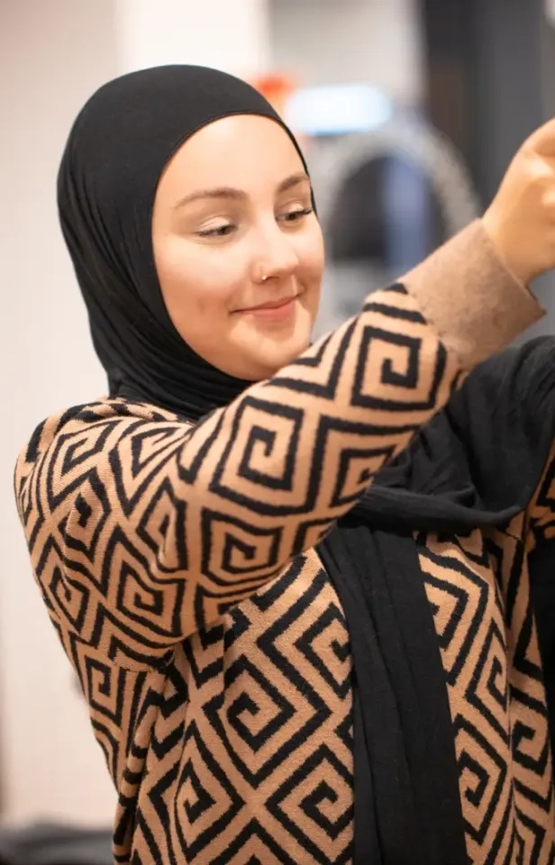 Eine Frau mit schwarzem Hijab und beigem Pullover mit geometrischem schwarzem Muster lächelt sanft und hält dabei etwas außer Sichtweite.
