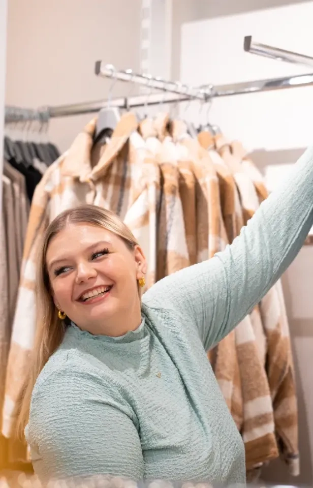 Eine lächelnde Frau in einem hellblauen strukturierten Oberteil greift nach oben zu einem Kleiderständer, im Hintergrund hängen beige und weiß karierte Jacken.