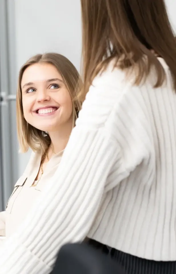 Eine junge Frau mit schulterlangem blondem Haar lächelt einer anderen Person in einem weißen Pullover, die von hinten zu sehen ist, strahlend zu. Die beiden scheinen sich drinnen freundlich zu unterhalten.