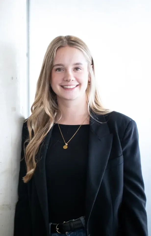 Eine junge Frau mit langen blonden Haaren, einem schwarzen Blazer und einem schwarzen Top lächelt in die Kamera. Sie trägt außerdem eine goldene Halskette und steht vor einem hellen Hintergrund.