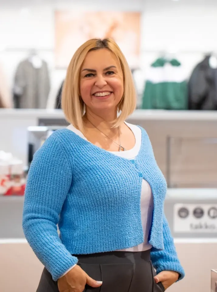 Eine lächelnde Frau mit glattem blondem Haar, die eine blaue Strickjacke über einem weißen Oberteil trägt, steht im Innenbereich vor verschwommenen Kleiderständern.