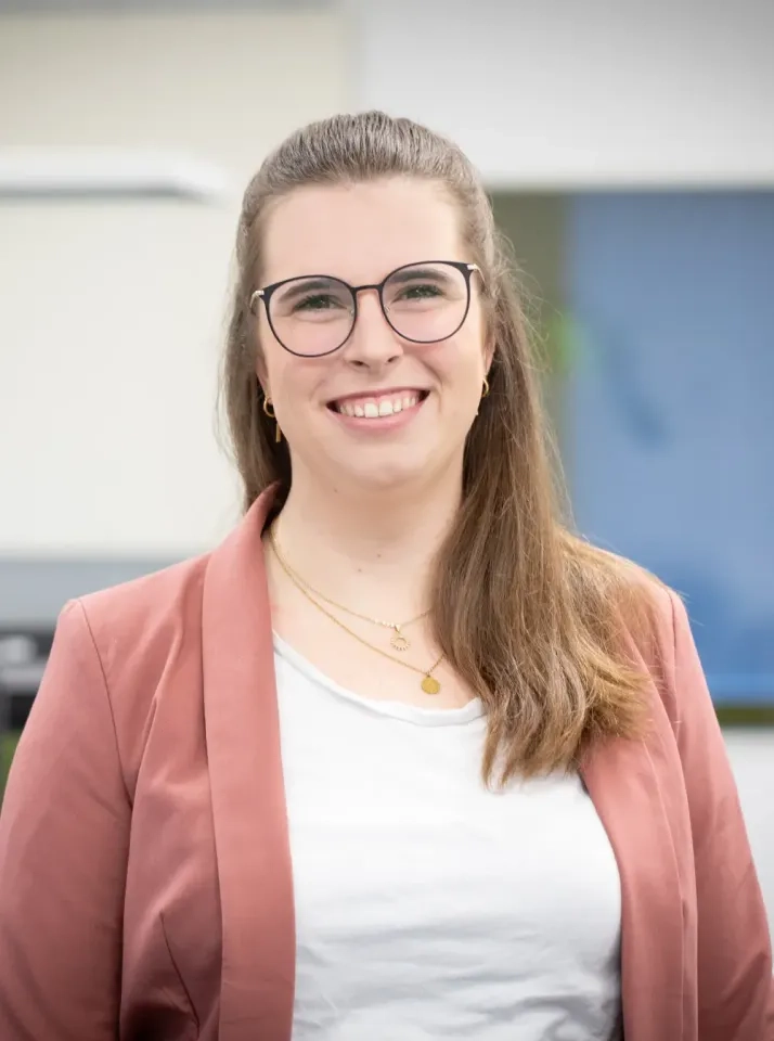 Eine Frau mit langen braunen Haaren, Brille und einem rosa Blazer über einem weißen Hemd lächelt in einem hell erleuchteten Innenbereich in die Kamera.