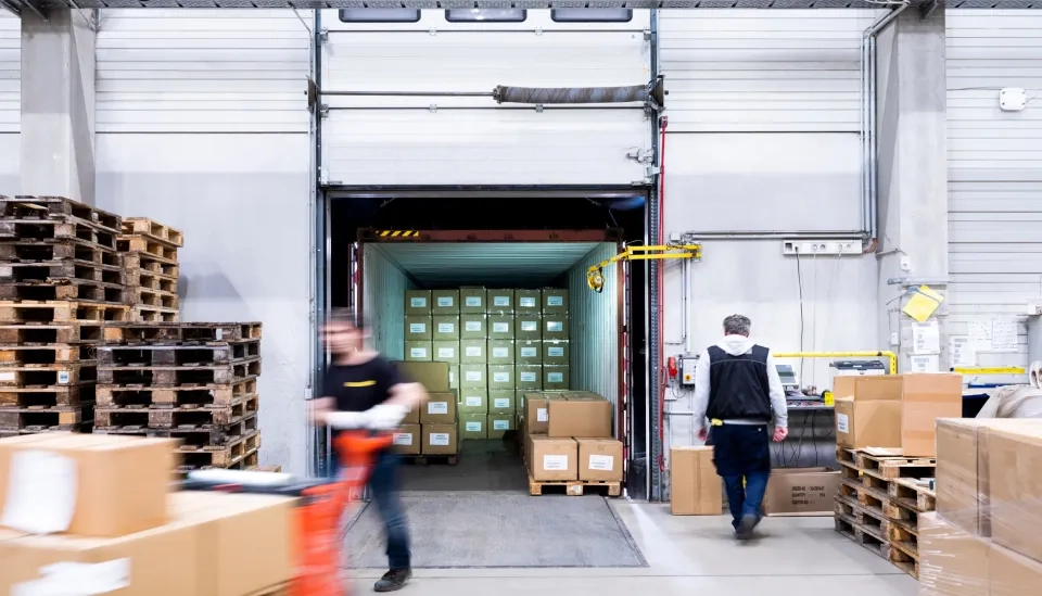 Zwei Arbeiter bewegen sich um eine mit Kisten gefüllte Laderampe in einem Lagerhaus. Ein LKW an der Rampe ist dicht mit gestapelten Kartons beladen. Holzpaletten und weitere Kisten liegen verstreut in der Nähe.