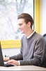 Ein junger Mann mit zurückgekämmtem braunem Haar, der einen grauen Pullover über einem weißen Hemd mit Kragen trägt, lächelt, während er an einem Schreibtisch in einem hellen, modernen Büro mit gelb eingefassten Fenstern auf einem Laptop tippt.