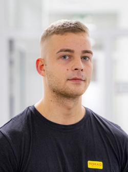 Ein junger Mann mit kurzen blonden Haaren und gestutztem Bart, der ein schwarzes T-Shirt mit einem gelben Takko-Logo trägt, steht in einem Innenbereich vor einem verschwommenen weißen Hintergrund.