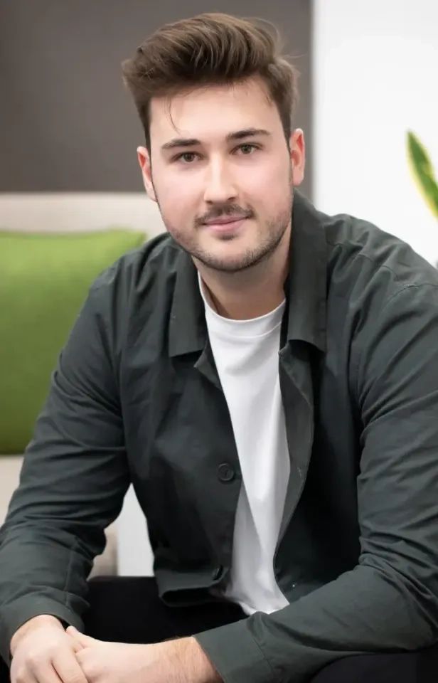 Ein junger Mann mit kurzen braunen Haaren und Bartstoppeln trägt ein weißes T-Shirt und eine dunkle Jacke, sitzt drinnen auf einem Sofa mit einem grünen Kissen, blickt in die Kamera und lächelt leicht.