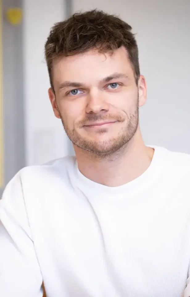 Ein junger Mann mit kurzen braunen Haaren und hellem Bart lächelt sanft. Er trägt einen weißen Pullover und sitzt drinnen vor einem unscharfen Hintergrund.