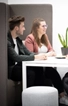 Zwei Personen sitzen nebeneinander an einem weißen Tisch in einer modernen Bürokabine, unterhalten sich und lächeln. Eine Topfpflanze steht auf dem Tisch, und eine neutral gefärbte Trennwand umgibt den Sitzbereich.