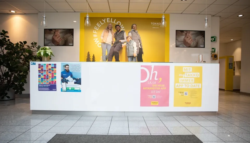 In einer hellen Lobby steht ein Rezeptionstresen mit bunten Plakaten an der Vorderseite. Dahinter befindet sich eine große gelbe Wand mit vier lächelnden, unterschiedlichen Menschen und den Aufschriften „#FEELYELLOW“ und „TAKKO FASHION“.