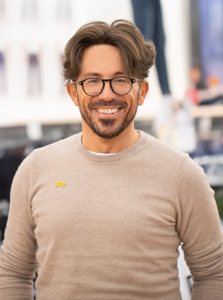 Ein lächelnder Mann mit Brille und welligem braunem Haar trägt einen beigen Pullover und eine kleine gelbe Anstecknadel und steht vor einem unscharfen Hintergrund im Innenbereich.