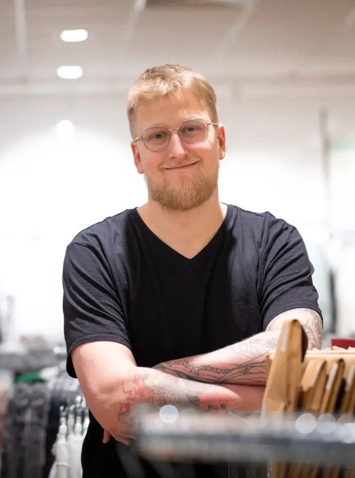 Ein lächelnder Mann mit kurzen blonden Haaren, Brille und Tattoos auf den Armen steht mit verschränkten Armen in einem hell erleuchteten Laden und trägt ein schwarzes T-Shirt mit V-Ausschnitt.