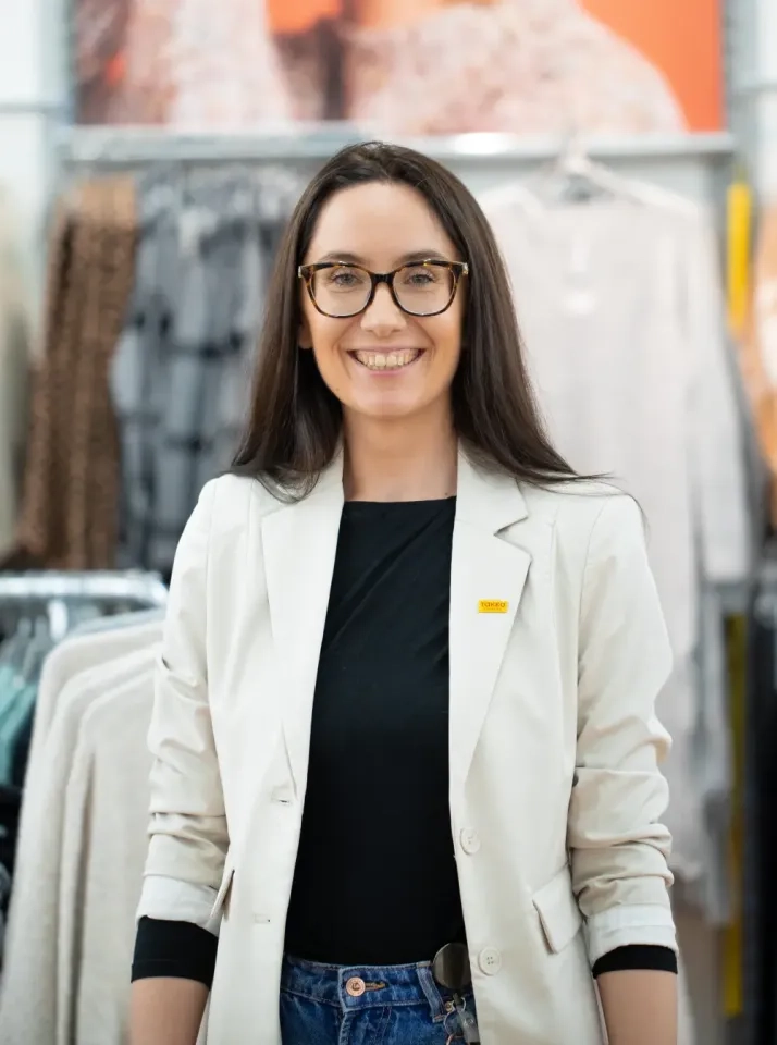 Eine Frau mit langen braunen Haaren und Brille steht lächelnd in einem Bekleidungsgeschäft. Sie trägt einen hellen Blazer über einem schwarzen Hemd und Jeans. Im Hintergrund sind Kleiderständer und verschwommene Kleidungsstücke zu sehen.
