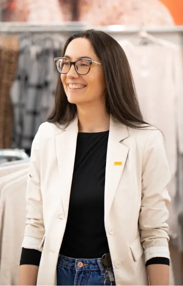 Eine lächelnde Frau mit langen braunen Haaren und Brille trägt einen cremefarbenen Blazer über einem schwarzen Top und Jeans. Sie steht im Innenbereich. Im unscharfen Hintergrund sind Kleiderständer zu sehen.