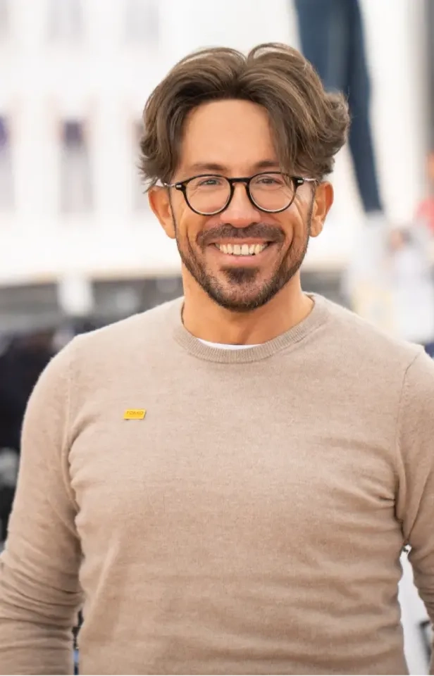 Ein lächelnder Mann mit Brille, welligem braunem Haar und einem beigen Pullover steht im Freien. Der Hintergrund ist leicht verschwommen und suggeriert eine städtische oder öffentliche Umgebung.
