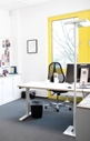 Ein helles Büro mit Schreibtisch, Computerbildschirmen, schwarzem Bürostuhl, Abfalleimer und gelb gerahmtem Fenster. An einer Wand stehen weiße Schränke, und eine Pinnwand mit Fotos und Notizen ist sichtbar.