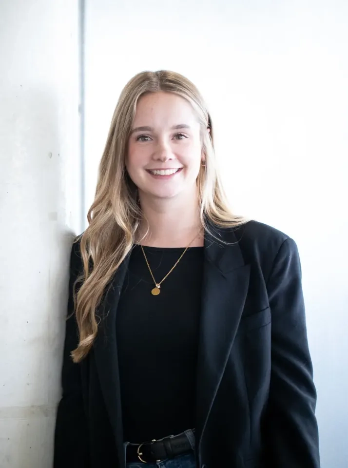 Eine junge Frau mit langen blonden Haaren, die einen schwarzen Blazer über einem schwarzen Oberteil und eine goldene Halskette trägt, steht lächelnd vor einem weißen Hintergrund.