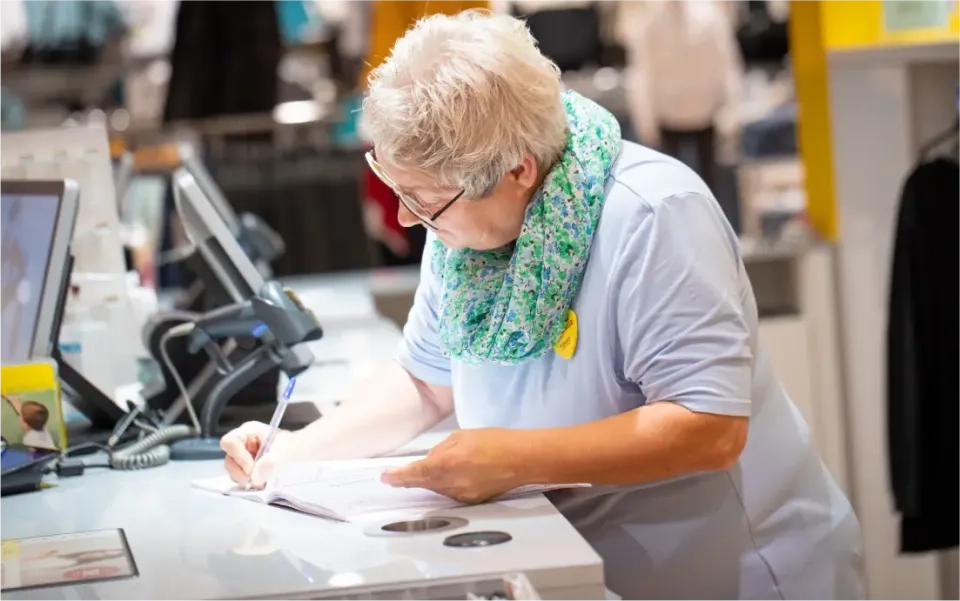 Eine ältere Frau mit kurzen grauen Haaren und Brille steht an einer Ladentheke und schreibt in ein Notizbuch. Sie trägt ein hellblaues Hemd und einen grün gemusterten Schal. Auf der Theke liegen ein Computer und ein Telefon.