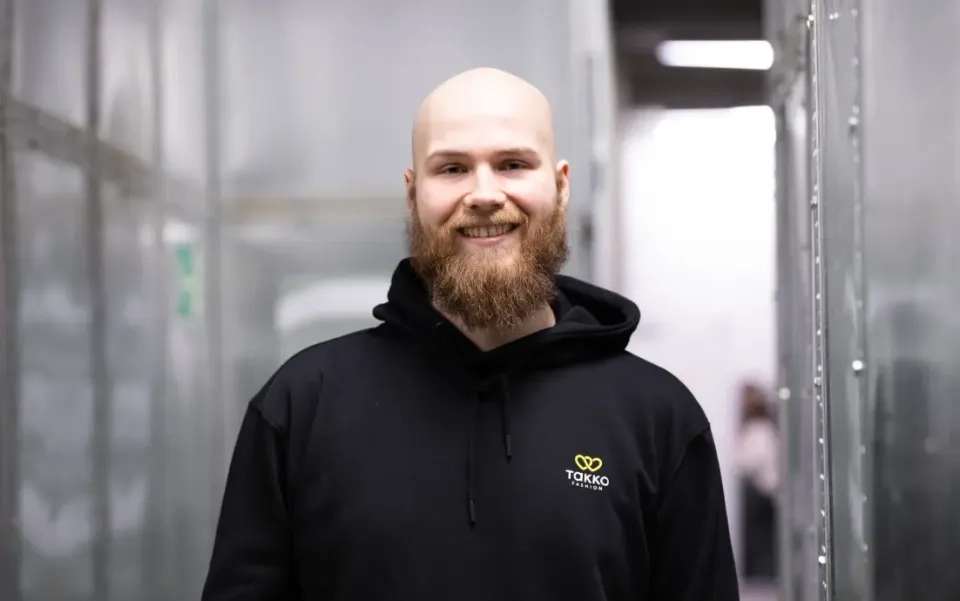 Ein lächelnder Mann mit Glatze und dichtem Bart trägt einen schwarzen Kapuzenpullover mit einem gelben TOKKO-Logo und steht in einem hellen, industriellen Flur mit metallischen Wänden.