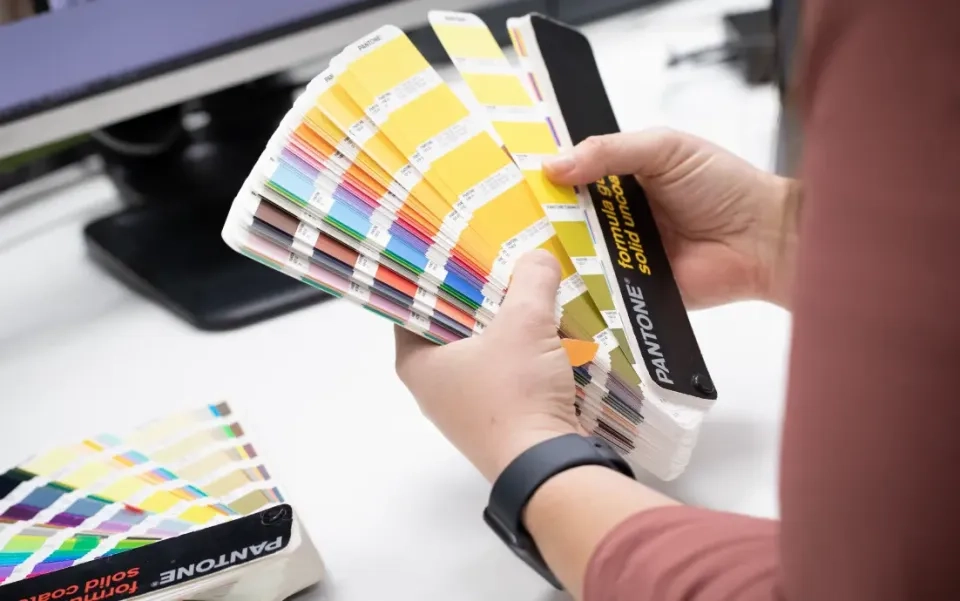 Eine Person hält einen Pantone-Farbführer in der Hand und blättert an einem Schreibtisch mit einem Computermonitor im Hintergrund durch gelbe Farbmuster.