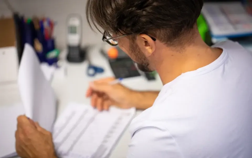 Ein Mann mit Brille und weißem Hemd sitzt an einem unordentlichen Schreibtisch, betrachtet Dokumente und schreibt mit einem Stift. Im Hintergrund sind Büromaterial, Papiere und ein Telefon zu sehen.