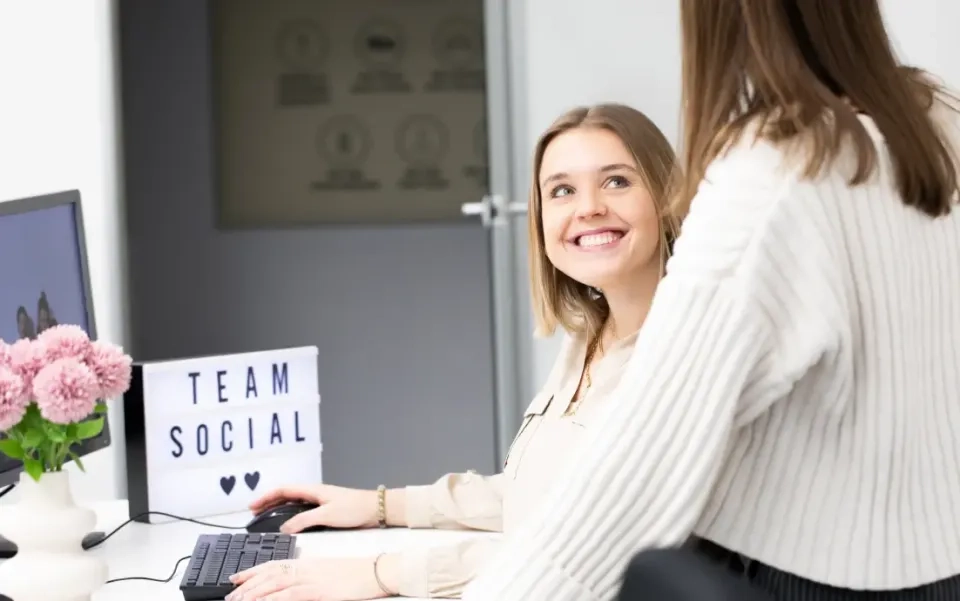 Zwei Frauen sitzen in einem Büro; die eine sitzt an einem Schreibtisch und lächelt die andere an. Auf dem Schreibtisch stehen ein Computer, Blumen und eine Leuchtbox mit der Aufschrift „TEAM SOCIAL“. Die Umgebung ist hell und modern.