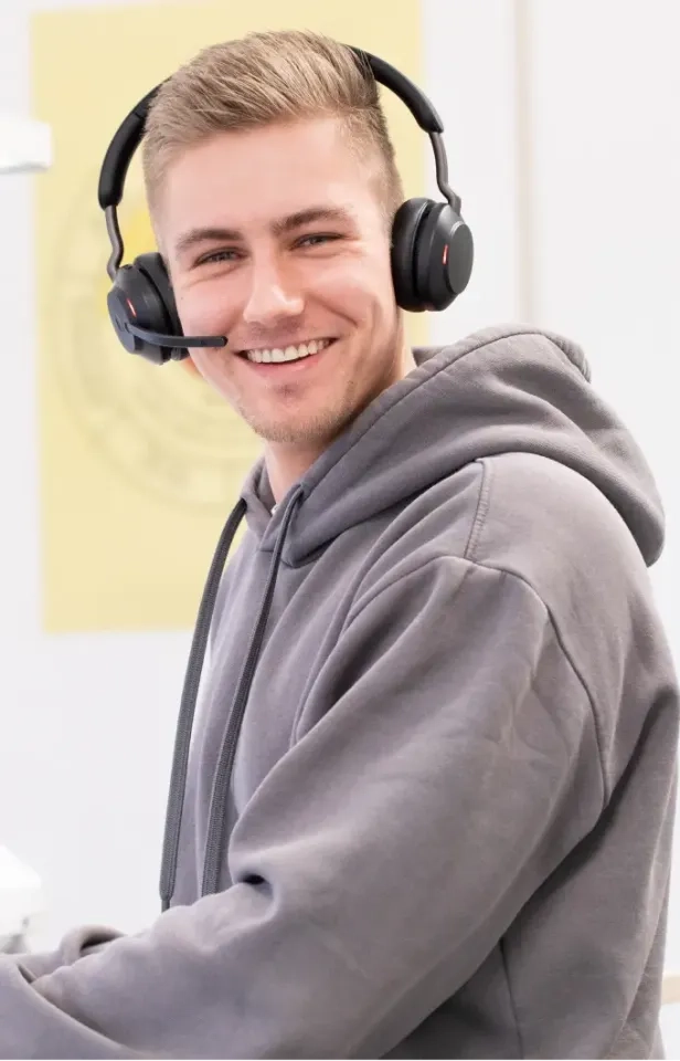 Ein junger Mann mit grauem Kapuzenpullover und schwarzem Headset mit Mikrofon sitzt lächelnd im Haus und deutet damit an, dass er sich unterhält oder im Kundendienst arbeitet.