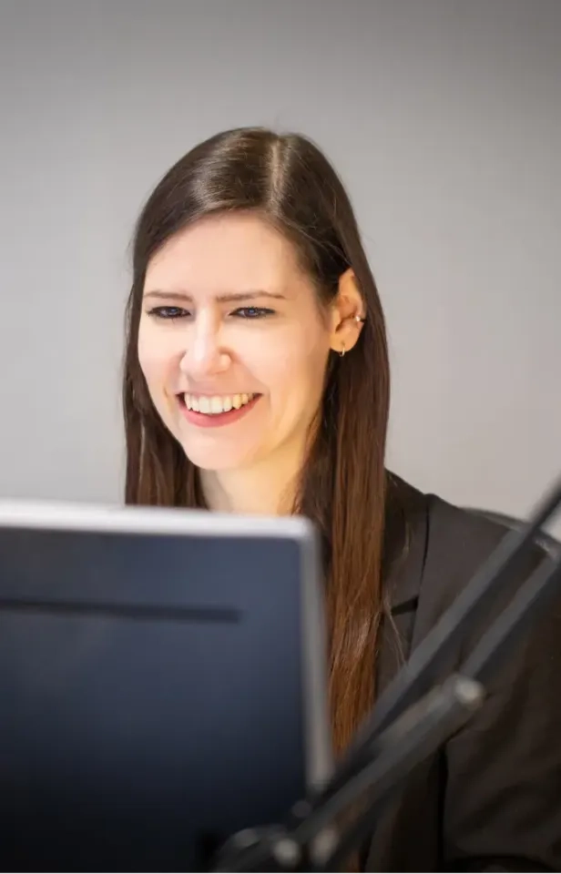 Eine Frau mit langen braunen Haaren und einem schwarzen Blazer lächelt, während sie auf einen Computermonitor blickt. Im Vordergrund ist ein Mikrofonarm zu sehen. Der Hintergrund ist schlicht und hell.