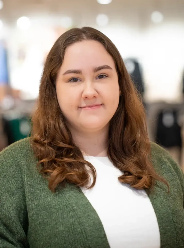 Eine junge Frau mit langem, welligem braunem Haar trägt eine grüne Strickjacke über einem weißen Oberteil und steht in einem Innenbereich vor einem unscharfen Hintergrund. Sie blickt in die Kamera und lächelt leicht.