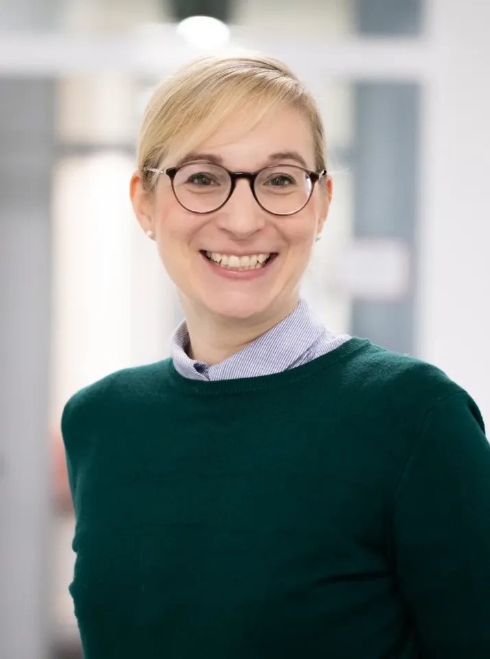 Eine Frau mit blondem Haar, Brille und einem grünen Pullover über einem Hemd mit Kragen lächelt in einem hellen Innenraum in die Kamera.