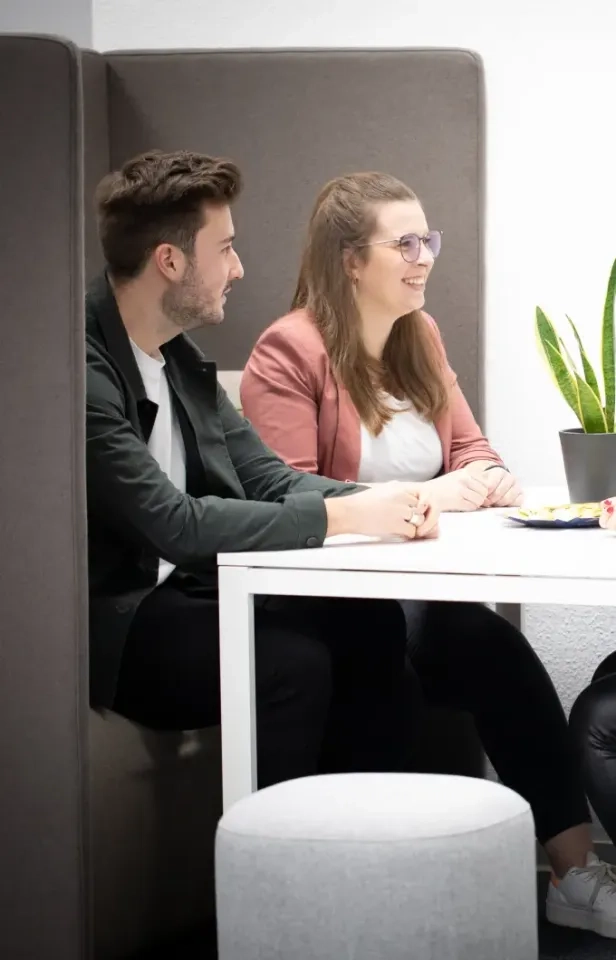 Zwei Personen sitzen nebeneinander an einem weißen Tisch in einer modernen Bürokabine und unterhalten sich. Auf dem Tisch stehen eine grüne Topfpflanze und ein Teller mit Snacks. Beide wirken freundlich und aufmerksam.
