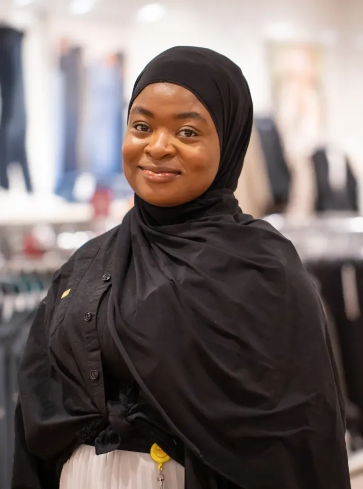 Eine Frau mit schwarzem Hijab und schwarzer Kleidung steht in einem Gebäude und lächelt sanft. Im Hintergrund sind Kleiderständer mit Kleidung verschwommen.