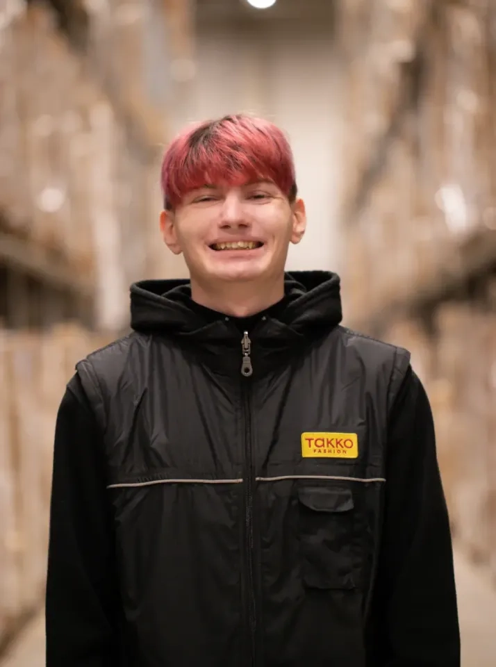 Eine junge Person mit kurzen, rosa und braunen Haaren lächelt in die Kamera. Sie trägt eine schwarze Weste mit einem gelben TAKKO-Logo und steht in einem Lagergang voller Regale und Kisten.