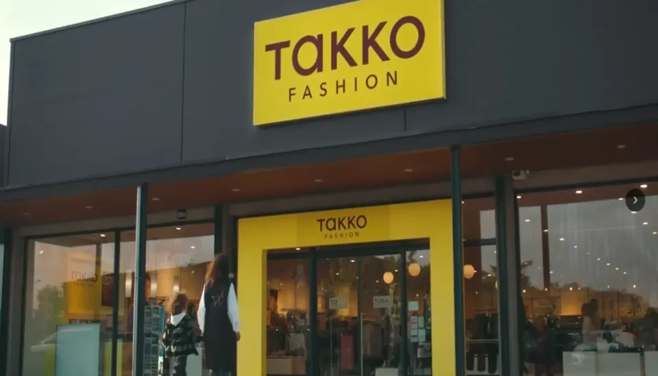 Das Bild zeigt den Eingang eines Takko Fashion Stores mit einem gelben Schild und großen Glastüren. Zwei Personen gehen auf den Eingang zu. Durch die Fenster ist das Innere des Ladens sichtbar.