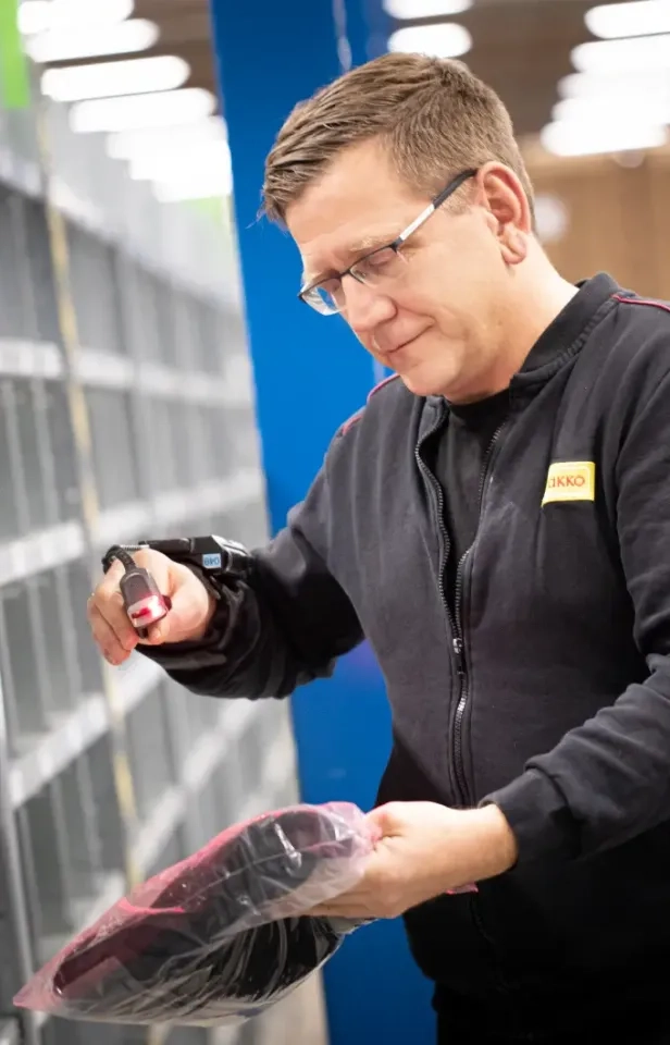 Ein Mann mit Brille scannt in einem Lagerhaus einen verpackten Artikel mit einem tragbaren Barcode-Scanner. Er trägt eine schwarze Jacke und steht neben Metallregalen.