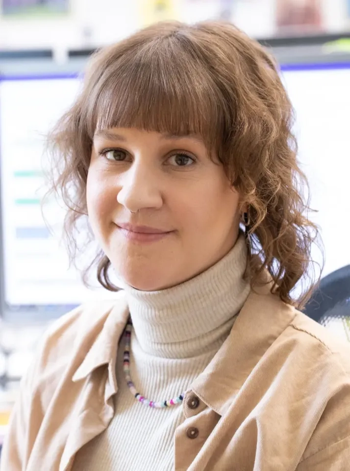 Eine Person mit welligem braunem Haar und Pony, die eine beige Jacke über einem hellen Rollkragenpullover und eine bunte Perlenkette trägt, sitzt im Innenbereich mit verschwommenen Computerbildschirmen im Hintergrund und lächelt sanft in die Kamera.