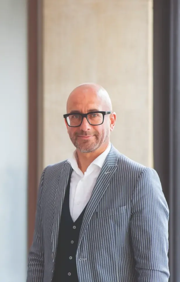 Ein Mann mit Glatze und Brille trägt ein weißes Hemd, eine schwarze Weste und einen grauen Nadelstreifenblazer. Er steht in einem Gebäude, lächelt leicht, vor dem verschwommenen Hintergrund heller Wände.