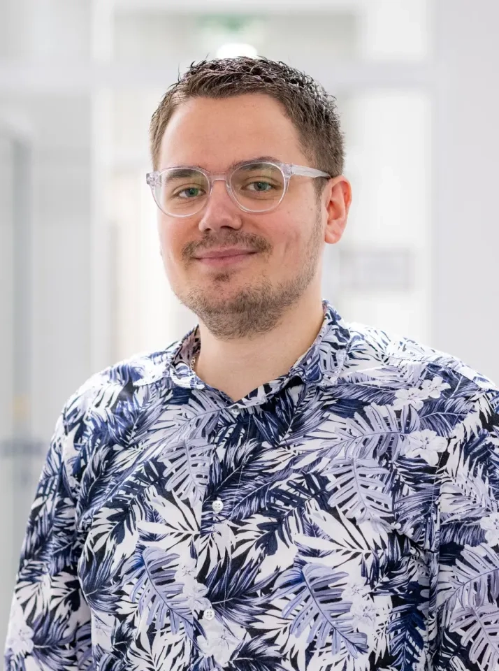 Ein Mann mit kurzen braunen Haaren, Brille und gestutztem Bart lächelt sanft. Er trägt ein Hemd mit blau-weißem Blattmuster. Der Hintergrund ist hell und leicht verschwommen, was auf eine Innenaufnahme schließen lässt.