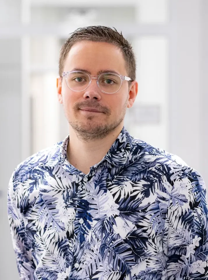 Ein Mann mit kurzen braunen Haaren, hellem Bart und Brille trägt ein blau-weiß gemustertes Hemd. Er steht in einem Raum vor einem hellen, unscharfen Hintergrund und blickt ruhig in die Kamera.
