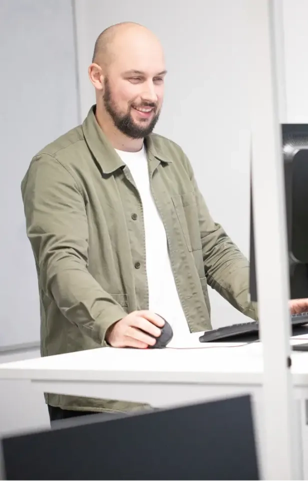 Ein Mann mit Bart und rasiertem Kopf, der eine grüne Jacke über einem weißen Hemd trägt, steht an einem Schreibtisch, bedient eine Computermaus und lächelt, während er auf den Monitor schaut.