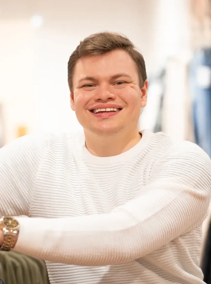 Ein junger Mann mit kurzen braunen Haaren, der einen weißen strukturierten Pullover und eine goldene Uhr trägt, lächelt vor einem leicht verschwommenen Innenhintergrund in die Kamera.
