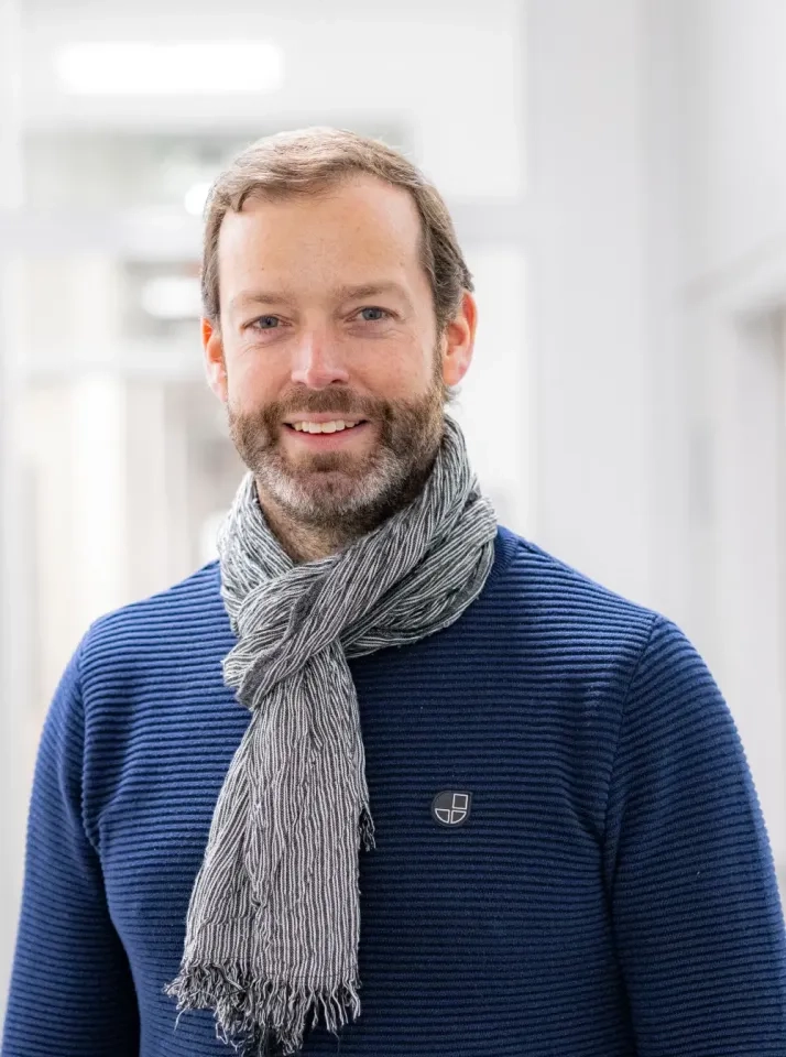 Ein Mann mit Bart und kurzen Haaren lächelt sanft. Er trägt einen marineblauen Strukturpullover und einen grauen Schal und steht in einem hell erleuchteten, modernen Raum mit weißen Wänden.