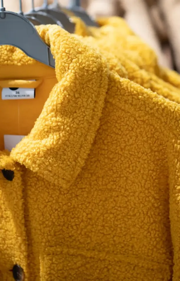 Nahaufnahme einer gelben, strukturierten Fleece- oder Sherpa-Jacke, die an einem Ständer hängt. Der Kragen und ein Teil des Etiketts im Inneren sind zu sehen. Der Stoff wirkt weich und gemütlich.